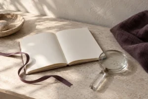 An open blank notebook with a plum ribbon and a glass magnifying lens on a stone desk.