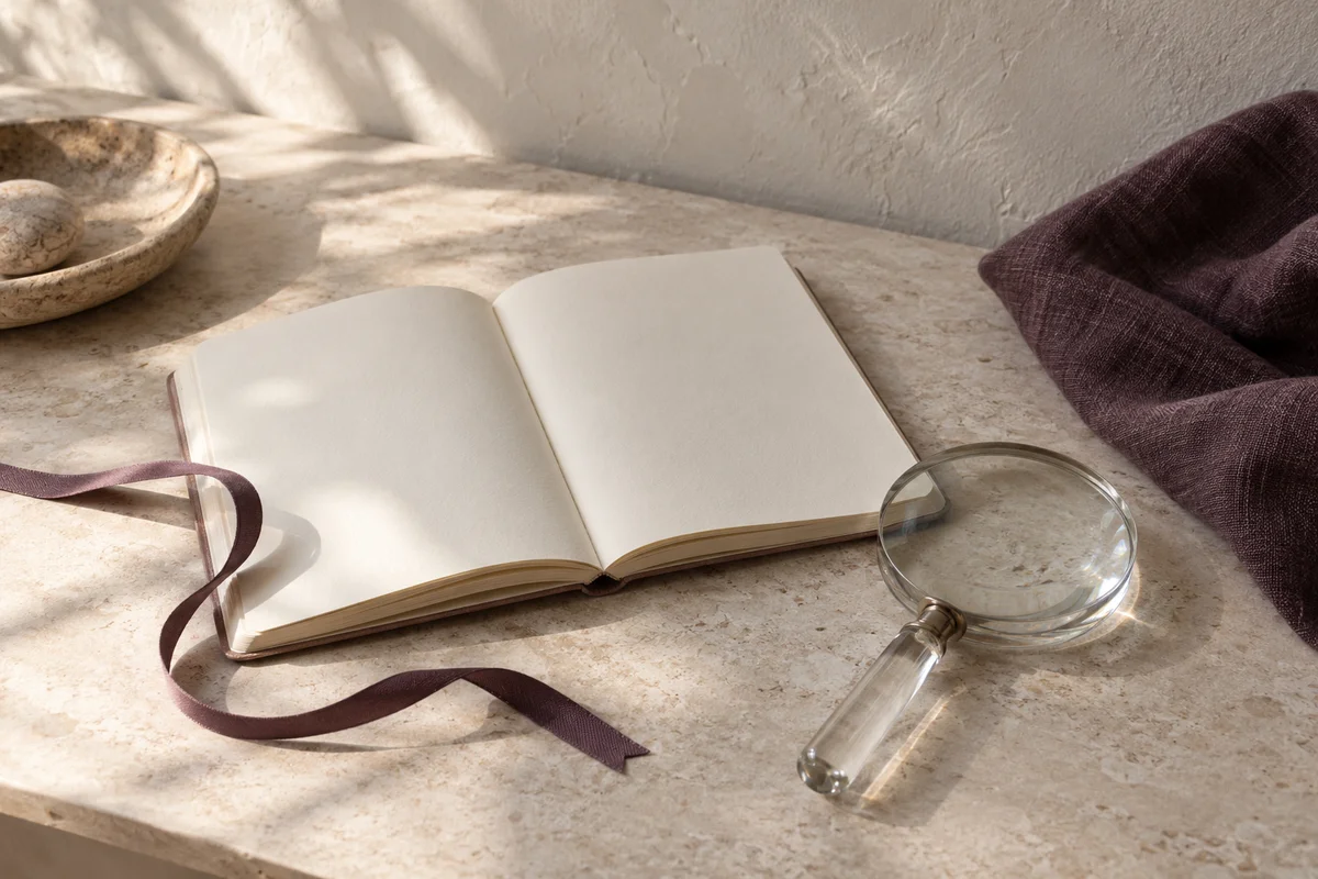An open blank notebook with a plum ribbon and a glass magnifying lens on a stone desk.